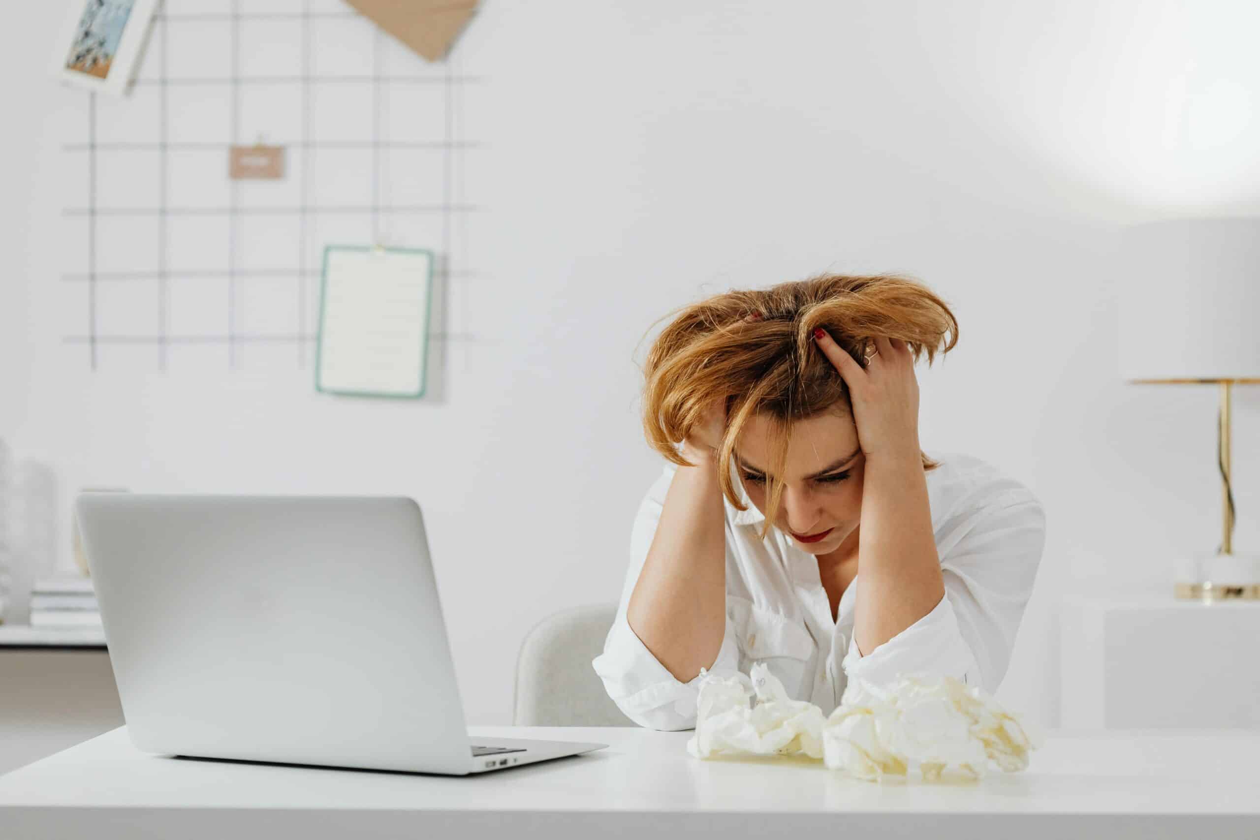 Frustrated woman in white shirt at desk with laptop, feeling overwhelmed.